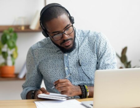 man sitting at desk wearing headphones taking notes whilst on a laptop